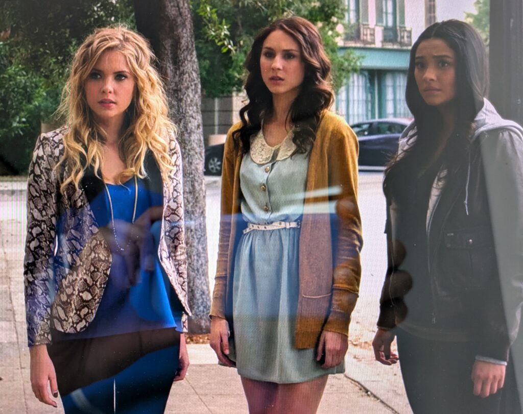 Hanna, Spencer, and Emily standing on a sidewalk looking concerned. Spencer is wearing light blue dress with a lace collar, silver buttons above the waist, and a thin white leather belt at the waist, along with a long burnt-orange cardigan