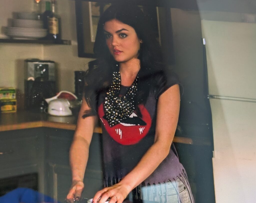 Aria in Ezra’s kitchen. She is wearing a dark gray t-shirt with a graphic of bright red lips. At the waist, the shirt is cut into strips of fringe that go past her gray jeans. She’s also wearing a sparkly black scarf around her neck
