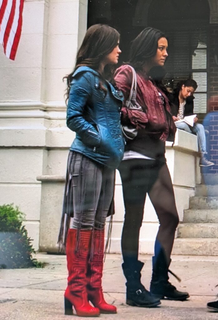 Wider shot of Aria in the same outfit; she is now wearing a blue leather jacket over the t-shirt, but we can see that the fringe goes down almost to her ankles, and that she is also wearing bright red knee-high heeled boots