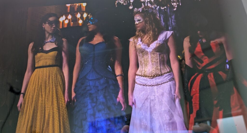 The Liars entering the masquerade ball together, all wearing floor-length gowns and masquerade-style masks. Spencer’s is mustard yellow with black accents; Emily’s is dark blue with a corset-style at the top and ruffles that look like fish scales from the waist down; Hanna’s is Renaissance style white and gold, which she is wearing along with a white flower crown-style tiara; and Aria’s is black and red alternating stripes, which she is wearing with elbow-length black gloves