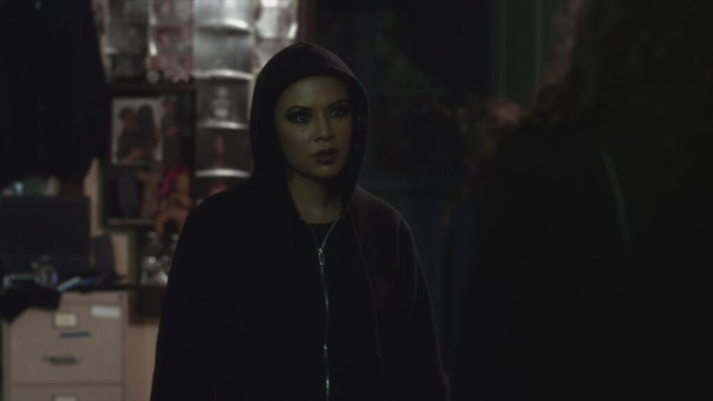Mona standing in a room at the Lost Woods Resort, looking angry. She is wearing a black hoodie with the hood up, while still in the dark eyeliner and lipstick she was wearing at the ball