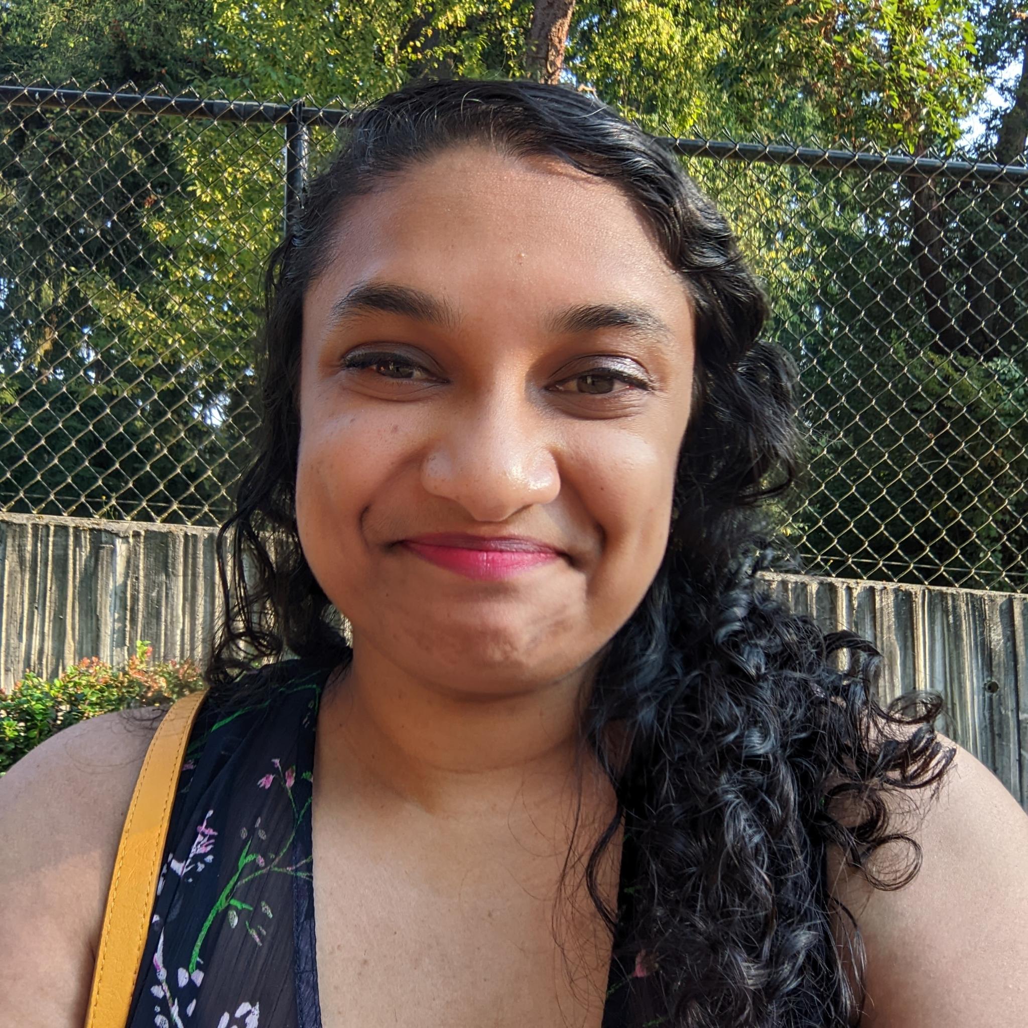 Headshot of a South Asian person with long curly black hair and light brown skin smirking at the camera. The photo is taken outside with trees and a fence visible in the background.