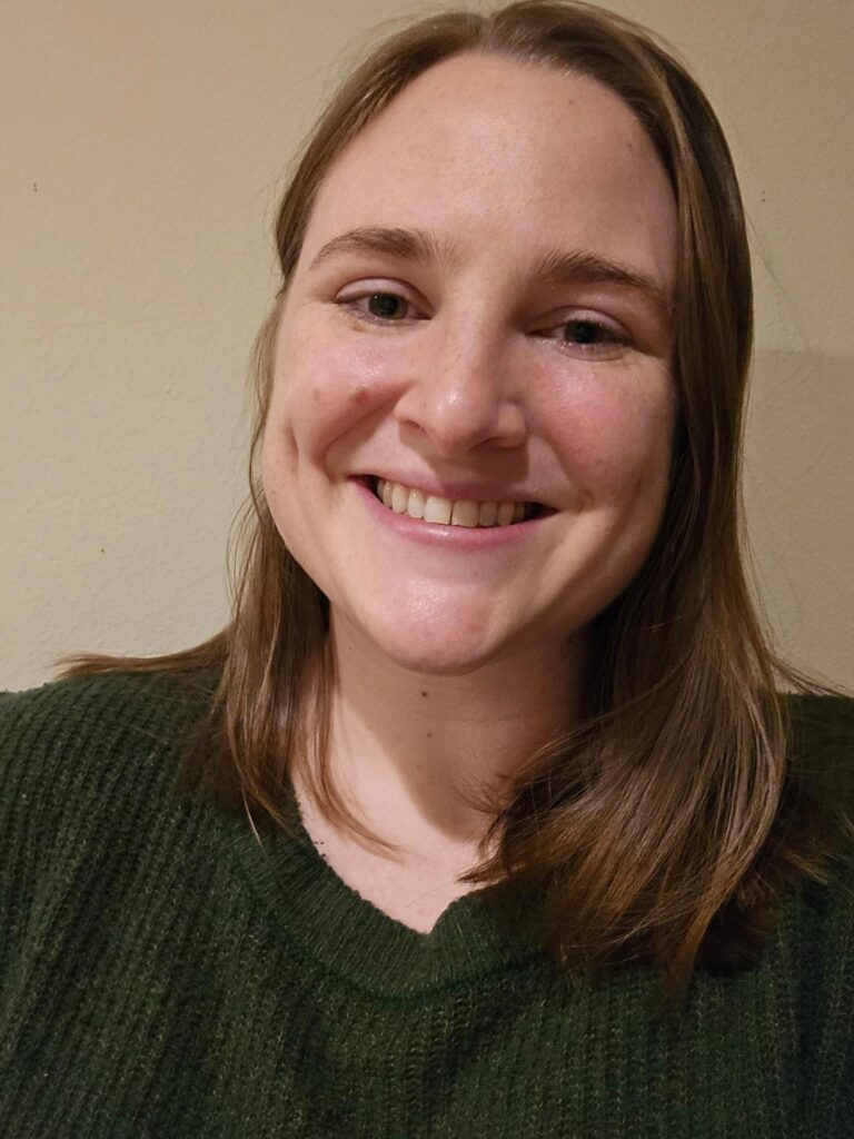 Headshot of a white woman with straight light brown shoulder-length hair wearing a green sweater and grinning at the camera