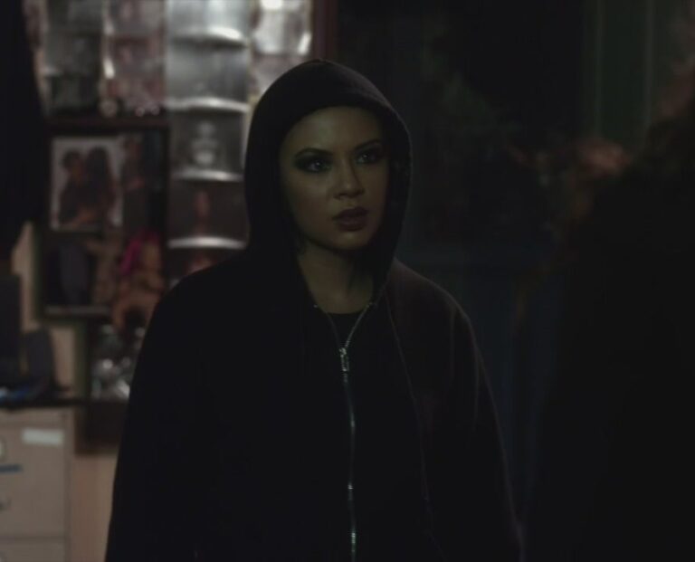 Mona standing in a room at the Lost Woods Resort, looking angry. She is wearing a black hoodie with the hood up, while still in the dark eyeliner and lipstick she was wearing at the ball