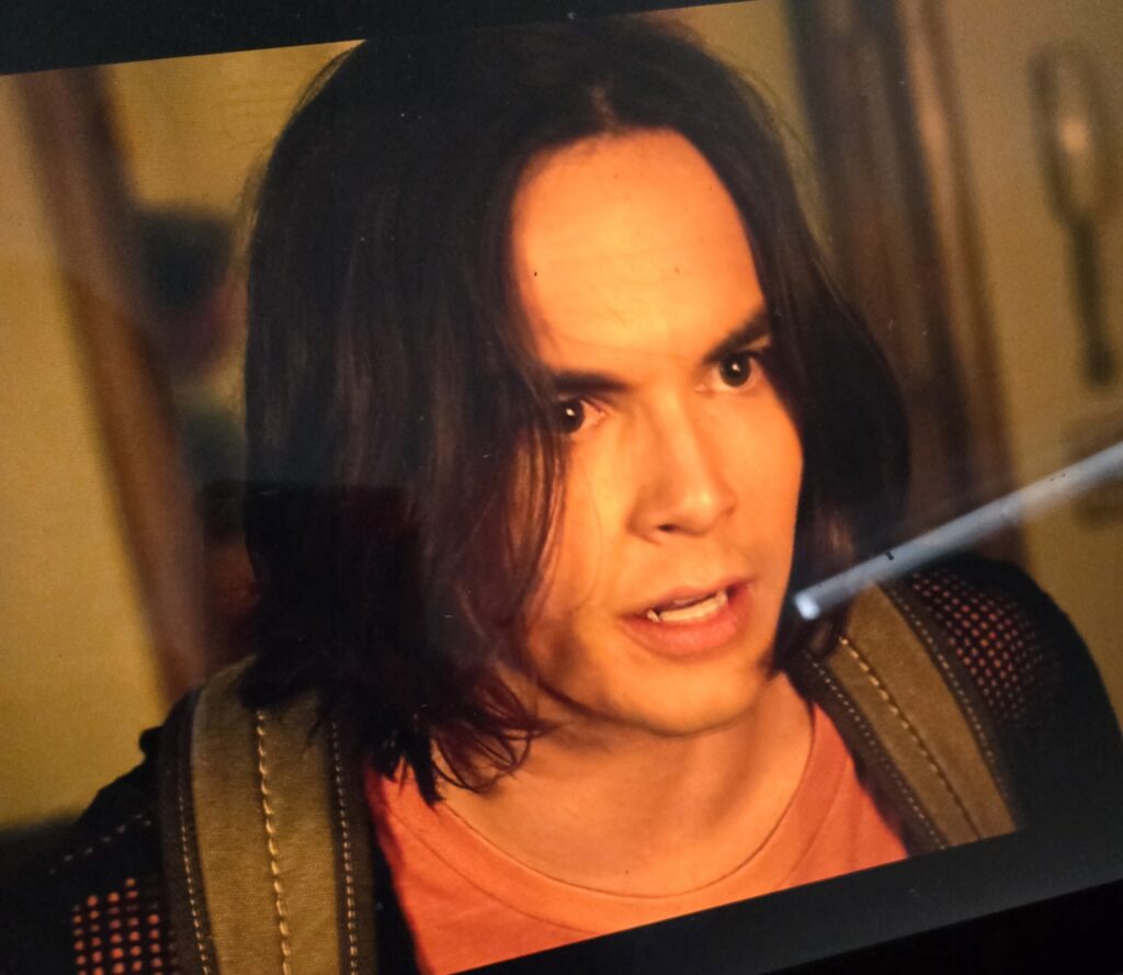 Caleb looks upset and is wearing a red shirt with the black mesh cardigan