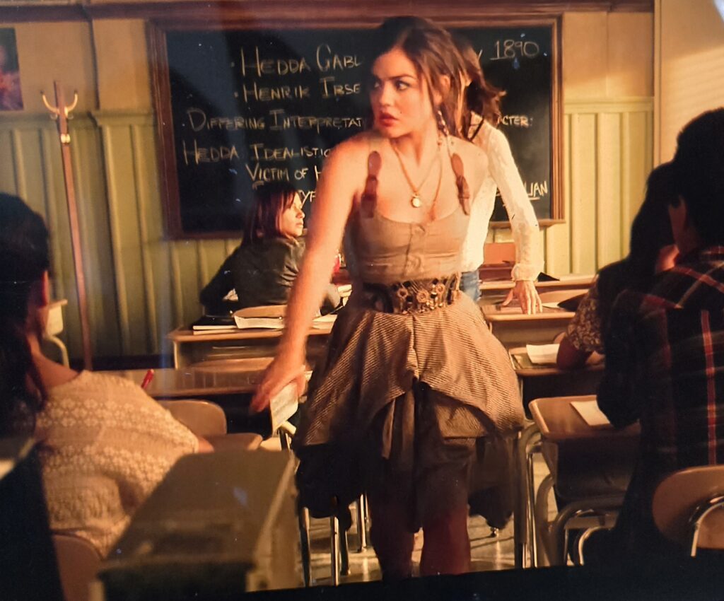 Aria is getting up from her desk in a dress that is striped with buckle straps and unfortunate layering, pleating and volume at the bottom