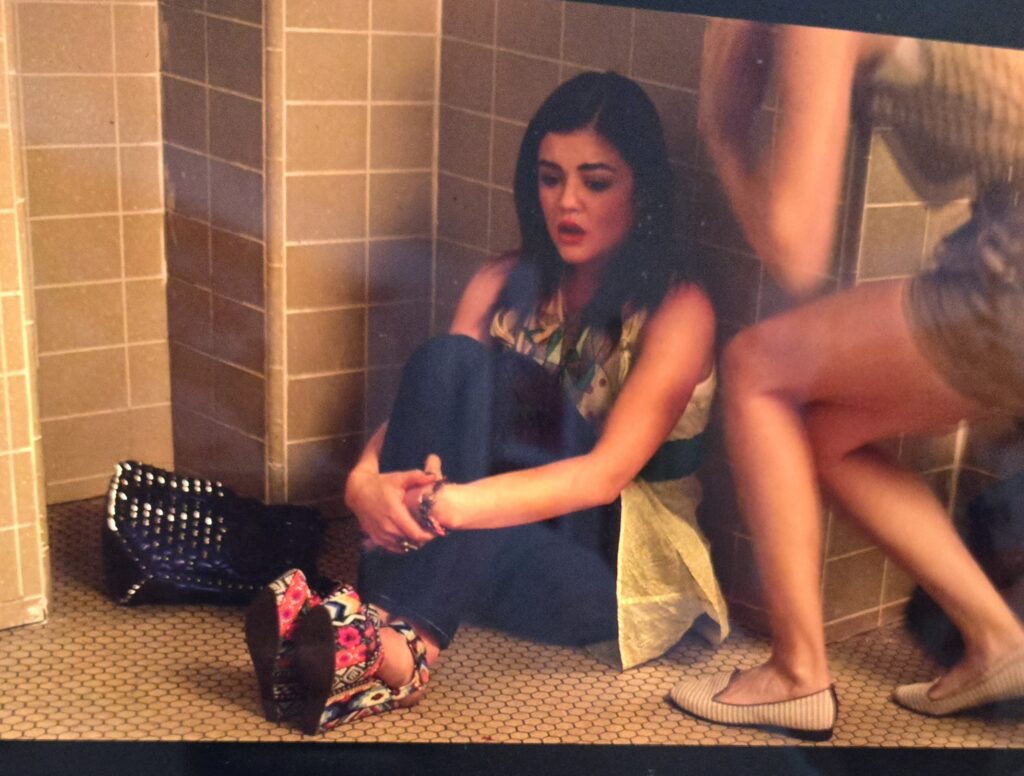 Aria crying in the bathroom. She is wearing a flowery scarf, a yellow sleeveless tunic-length top, blue jeans, and platform heels with a multi-colored flowery pattern