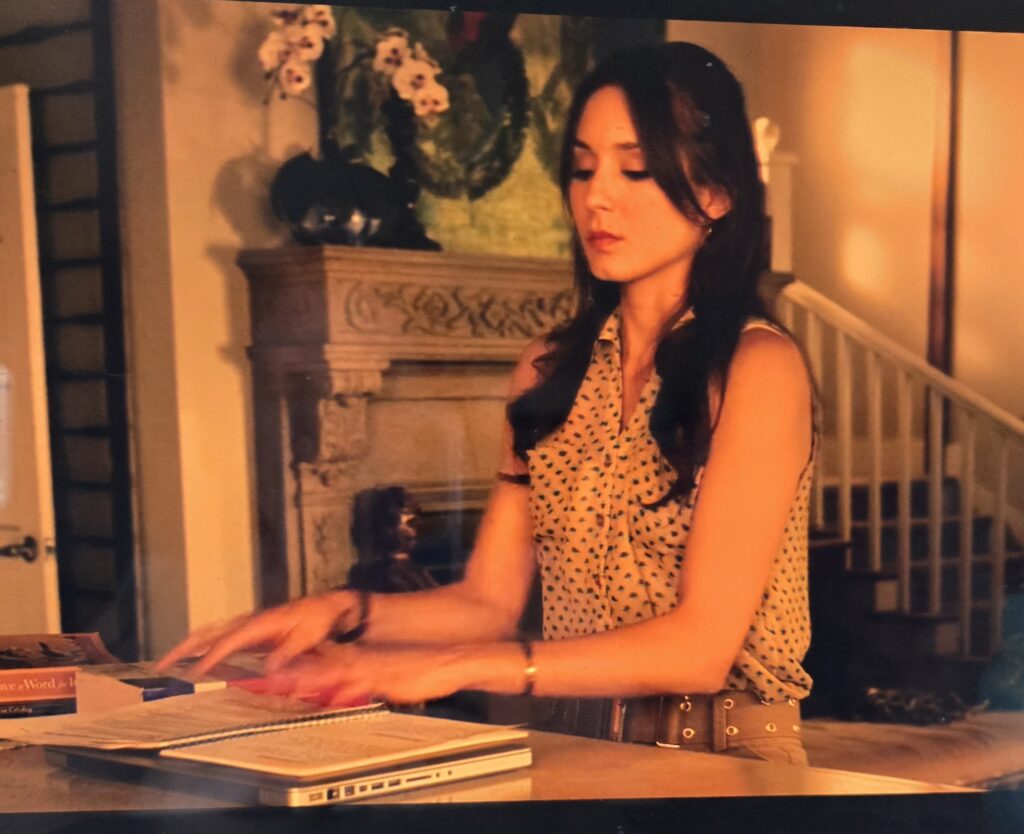 Spencer at home looking at a notebook. She is wearing a sleeveless white button-down with polka dots