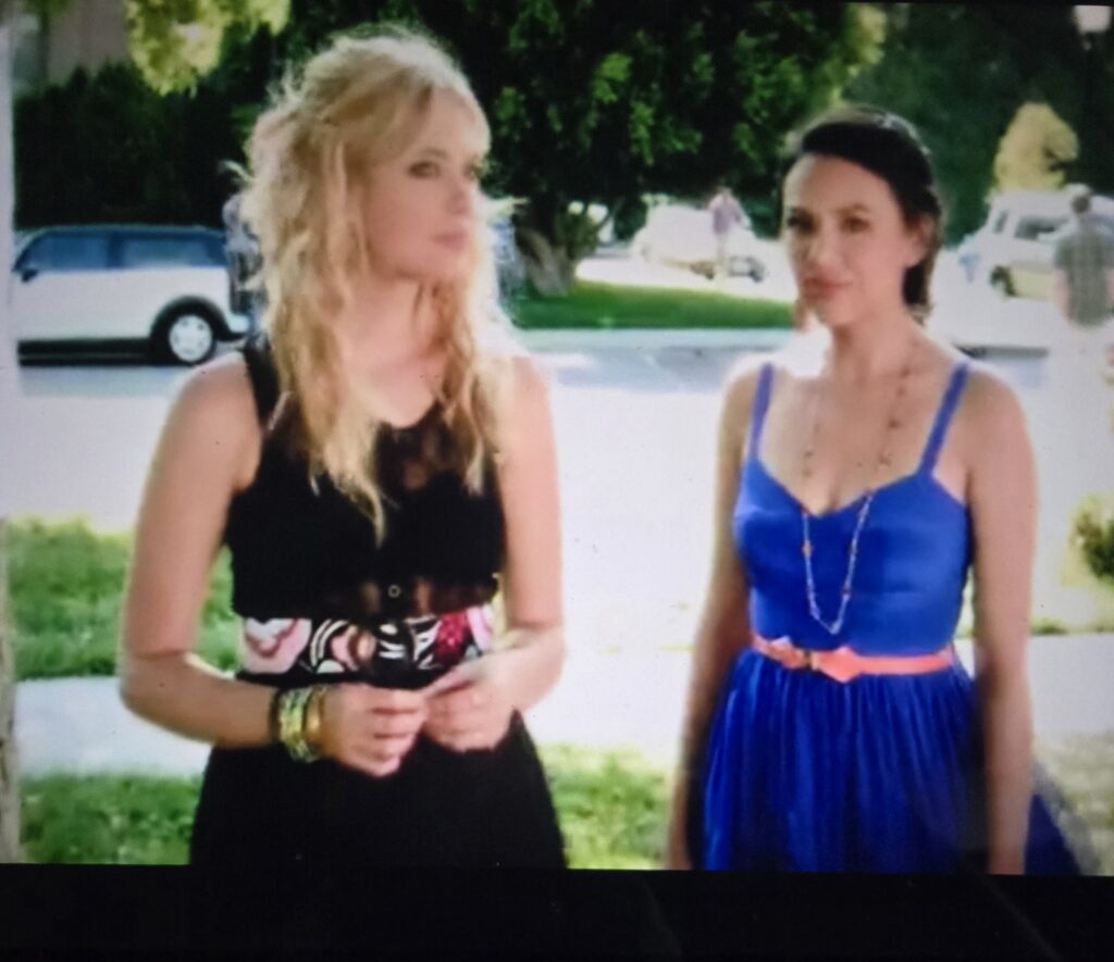 In a flashback scene - Mona in a blue dress and pink belt standing talking to Hanna