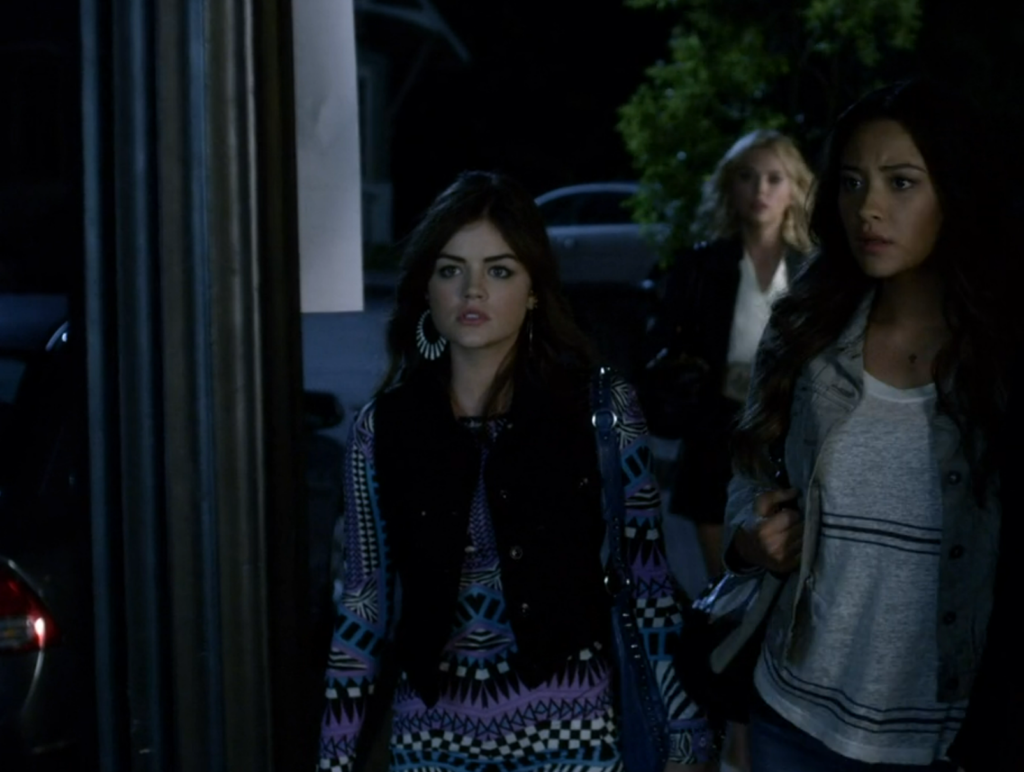 Aria on the sidewalk with the other Liars, wearing large hoop earrings and a long-sleeved dress with an intricate asymmetrical geometric pattern in black, lilac, white, and blue, topped with a dark gray jean vest