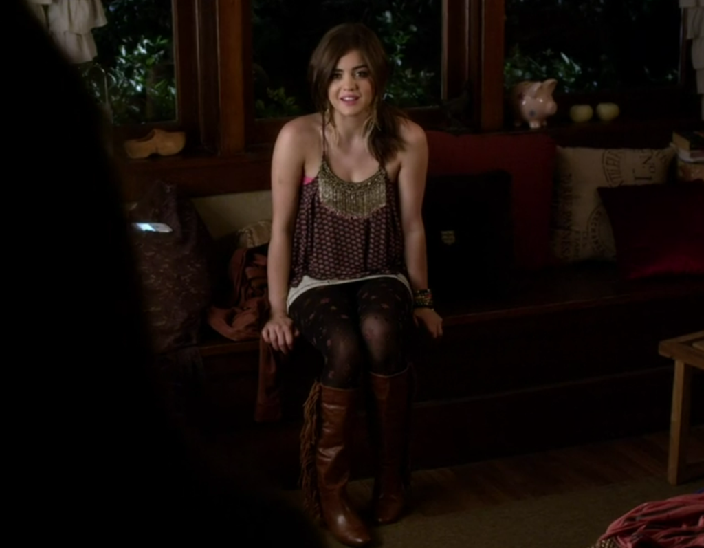 Aria sitting on her couch wearing a tank top that has a large metal necklace on the hem, along with patterned black tights and brown boots with fringe