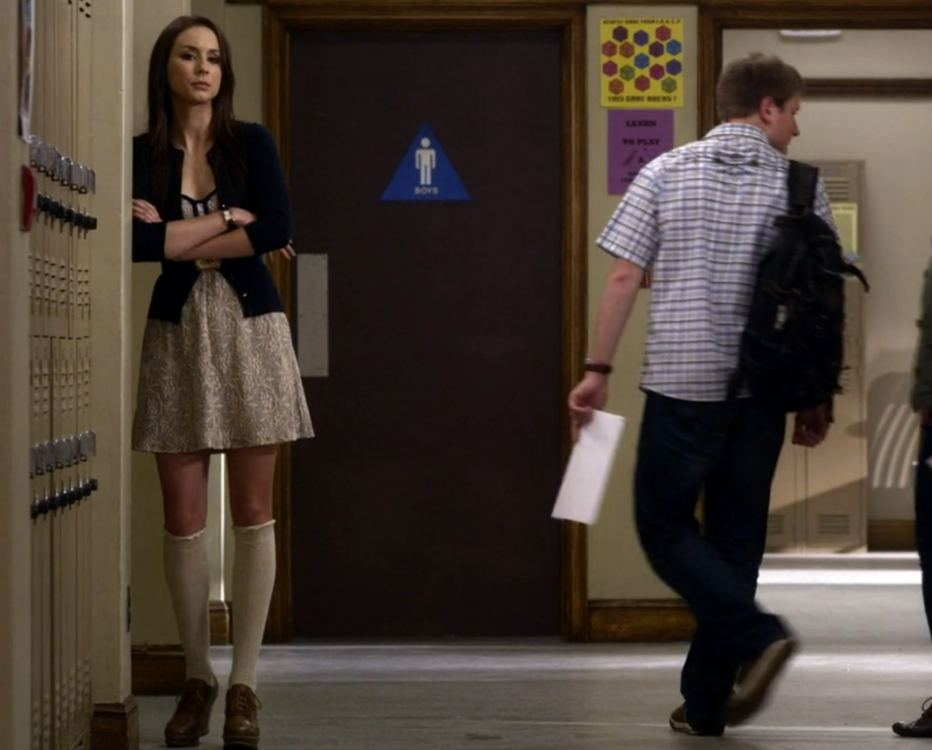 Spencer leaning on the lockers in the school hallway with her arms crossed over her chest, wearing a black blazer, over a cream colored dress, white knee-high socks, and brown oxford platforms.