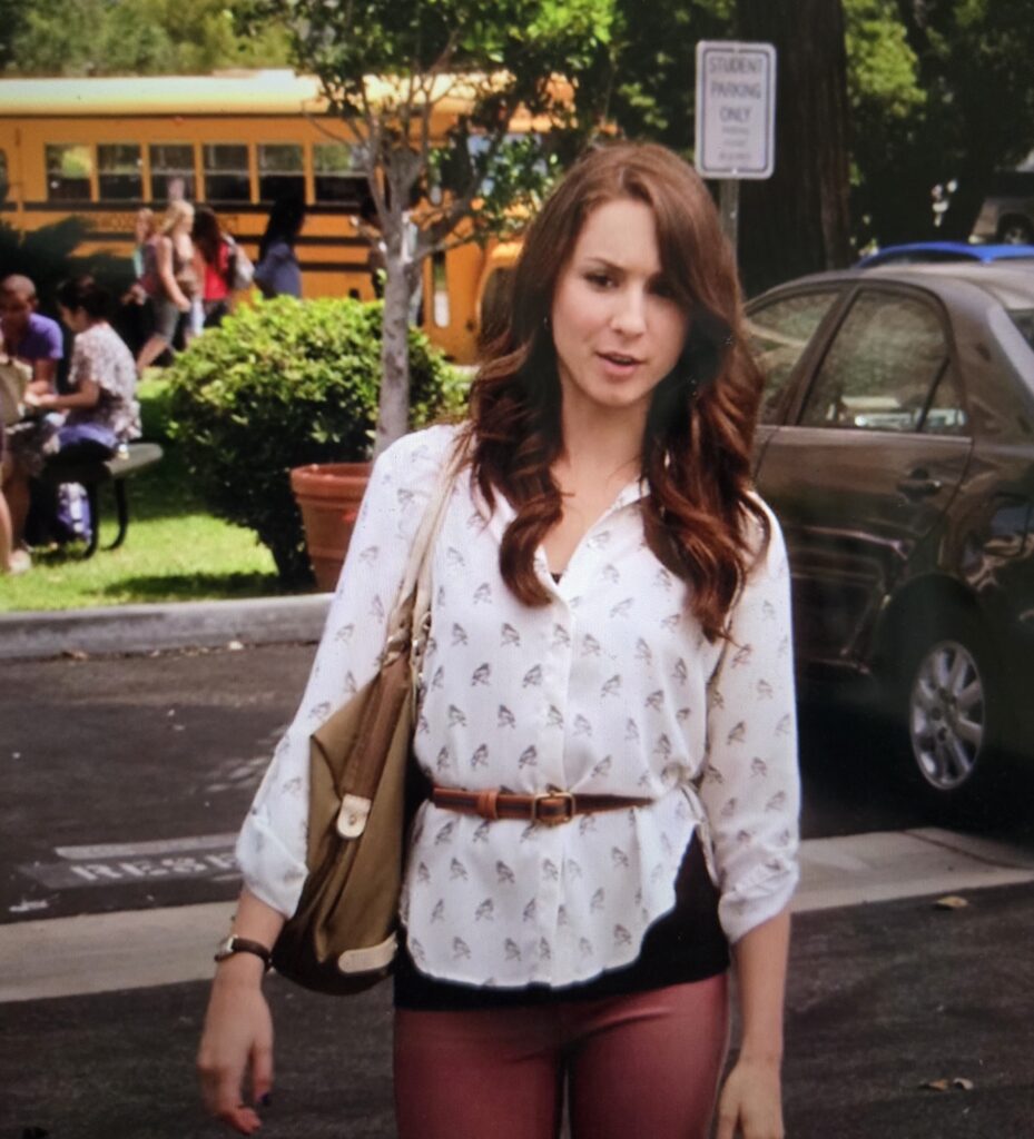 Spencer walking in a parking lot in a belted white shirt with (surprise!) birds on it. She is wearing her hair down and has a bag over her shoulder.