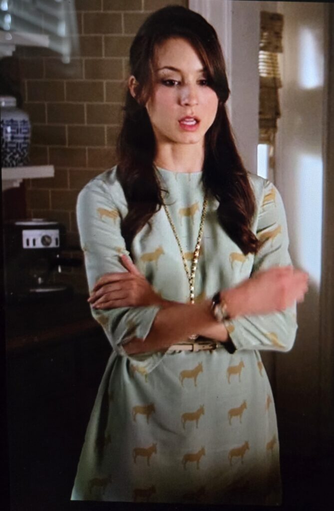 Spencer with arms crossed and looking down. She is wearing a light blue dress with brown donkeys on it with a white necklace.