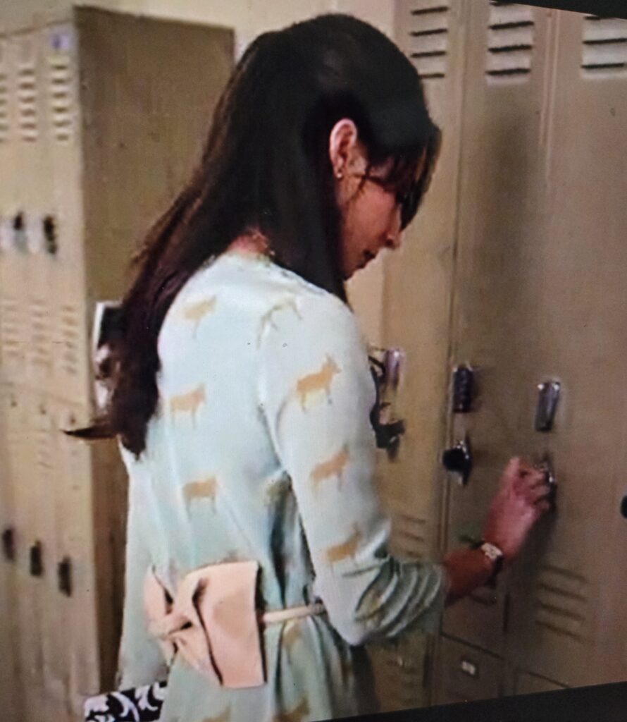 Spencer at her locker. The ugly giant bow on the back of her belt she is wearing over the light blue dress with brown donkeys is visible