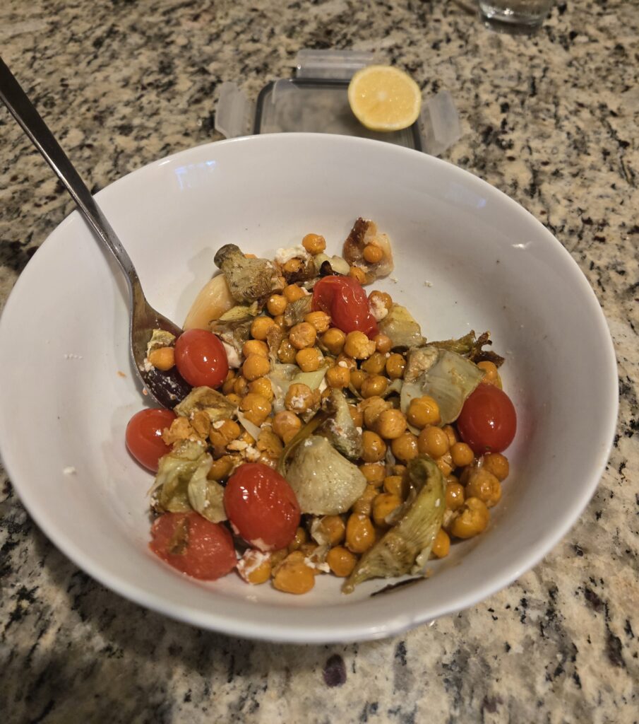 A bowl with a dish in it that has chickpeas, red cherry tomatoes, artichoke hearts, and feta cheese.