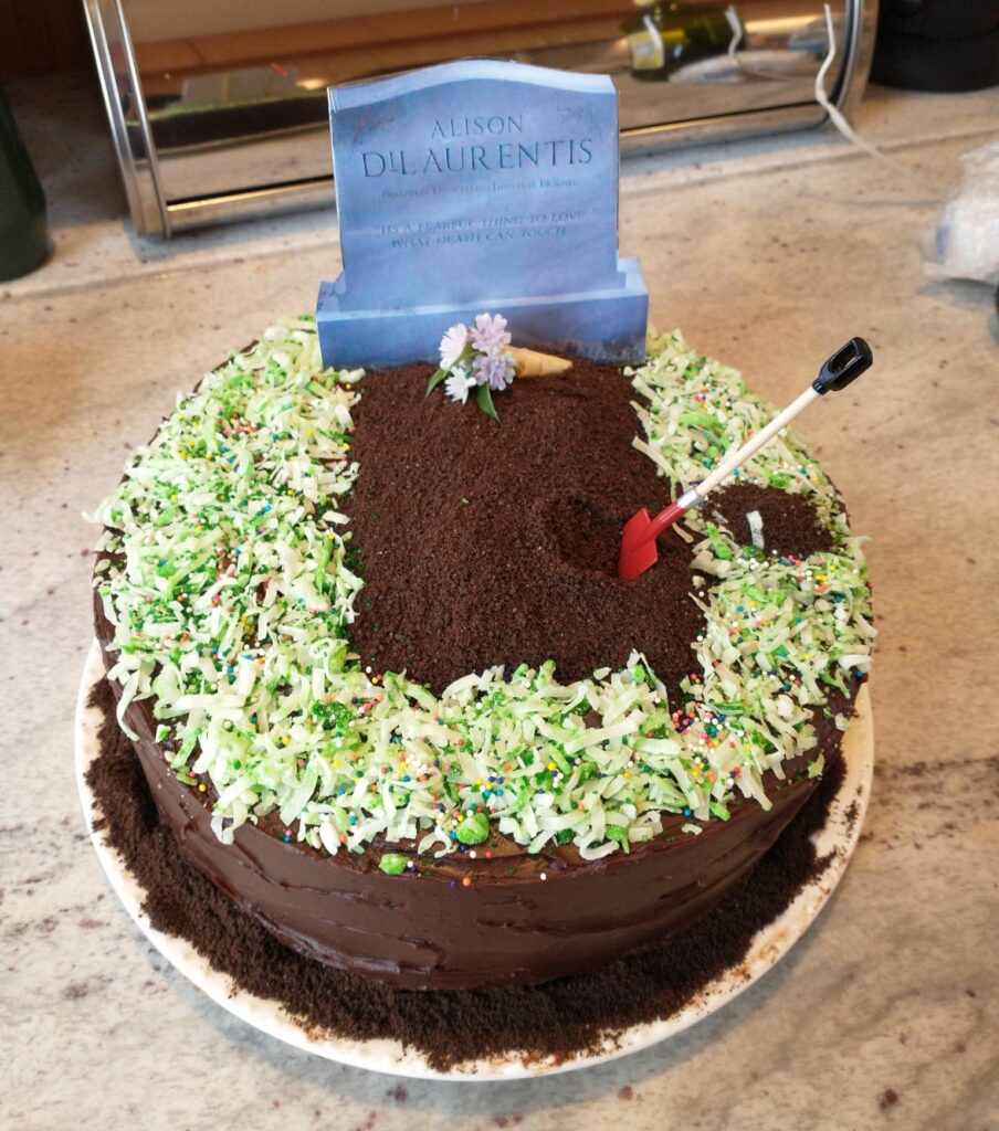 A beautiful chocolate cake with a replica of Ali’s grave on it. Some flowers are placed on the grave and there is a red shovel stuck in the cake. There are also green coconut flakes around the cake to look like grass. KC is a genius
