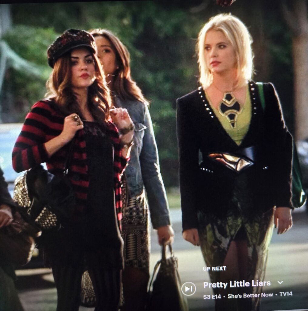 The Liars are walking on the sidewalk. Aria is wearing a horrible black hat with a red pattern and is making a pouty face. She has a red and black striped sweater over a black dress and black tights. Hanna is wearing a necklace that is so big and metallic and Black. It is kind of shaped like a spaceship. She also has a horrible black metal belt on and a weird asymmetrical green skirt