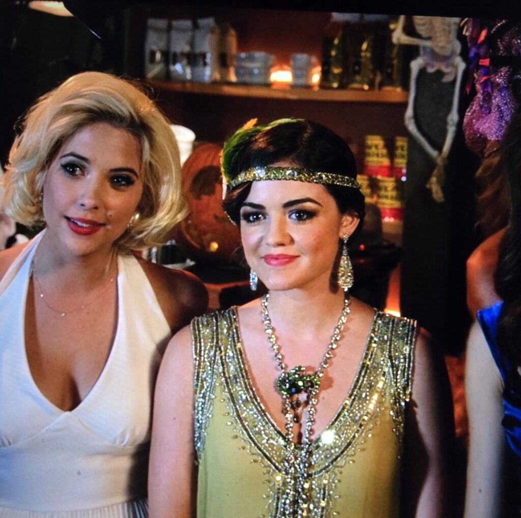 Aria standing with Hanna - Aria is dressed as Daisy from the Great Gatsby with a light green flapper dress and matching headband and a pretty giant silver earrings and necklace with big green jewels